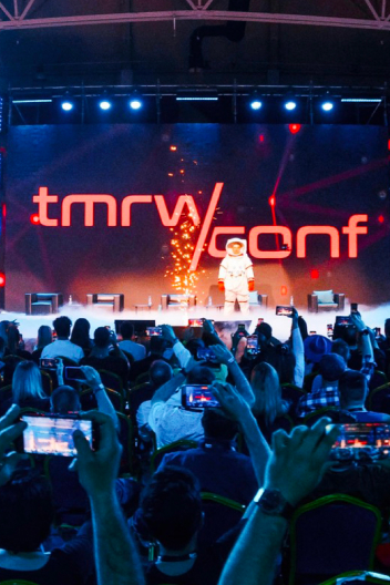 A crowd at a conference captures photos of a speaker onstage with the tmrw conf logo displayed prominently in the background.