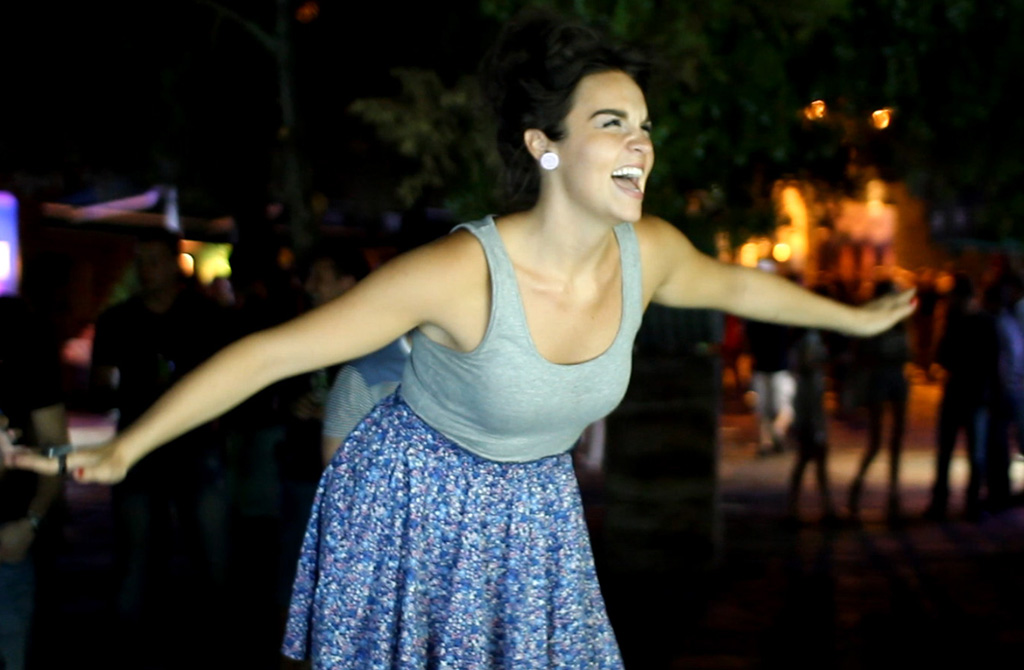 A joyful woman with her arms outstretched, spinning around at an outdoor nighttime event. The background is softly lit and blurred, highlighting her smiling expression and the motion of her twirling skirt.