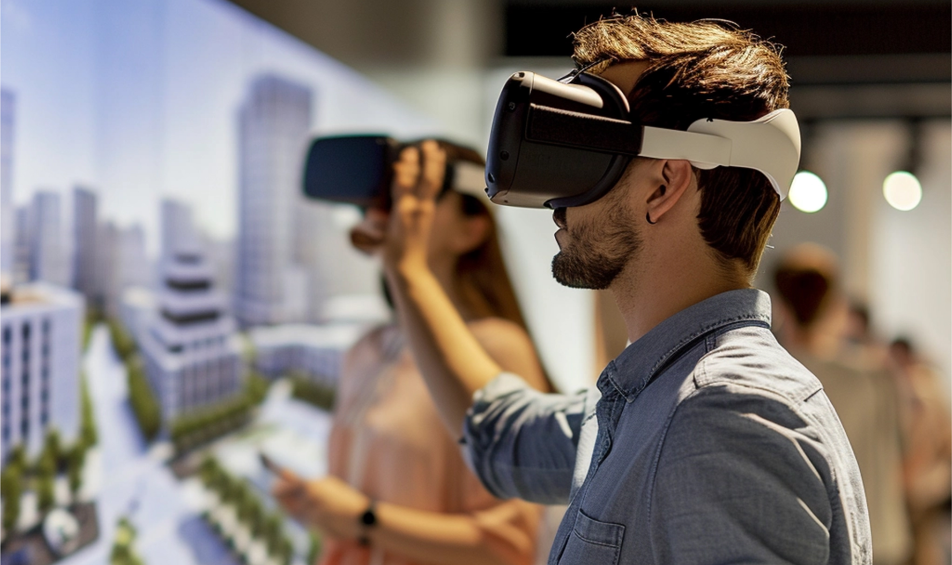 Muškarac i žena u ležernoj odeći stoje u zatvorenom prostoru i koriste VR naočare, sa zamućenim slikama modela gradskog pejzaža u pozadini, što sugeriše virtuelnu interakciju sa trodimenzionalnim okruženjem.