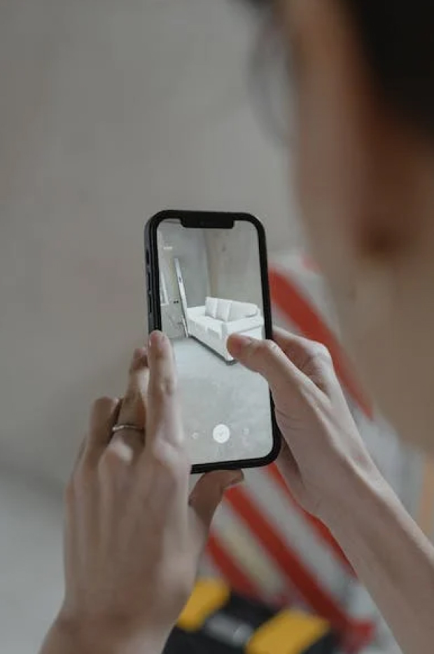 Ruka osobe koja drži pametni telefon, prikazuje model proširene stvarnosti (AR) bele sofe u prostoriji, demonstrirajući upotrebu AR-a u dizajnu enterijera.