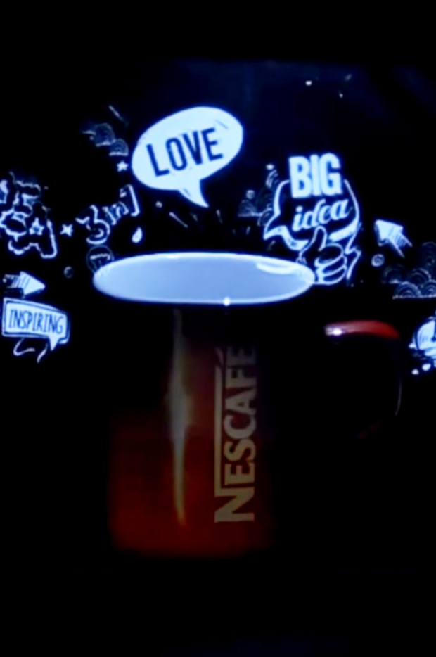 Krupni kadar šoljice za kafu sa animiranim crtežima i rečima poput 'LOVE' i 'BIG idea' projektovanim na nju, ilustrujući kreativno oglašavanje ili digitalnu umetnost.