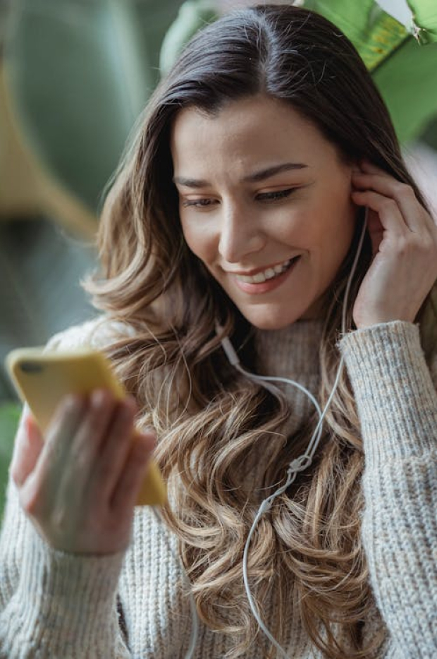 Nasmejana žena sa slušalicama gleda u svoj pametni telefon, njen izraz lica odražava uživanje i angažovanost u sadržaju koji gleda.