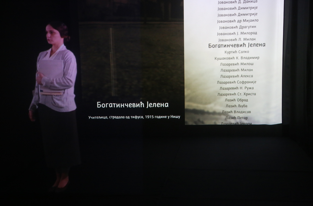Holografska projekcija istorijske ličnosti u prirodnoj veličini, odevene u periodičnu odeću, koja stoji pored zidne liste imena, deo obrazovne izložbe ili memorijalne prezentacije.