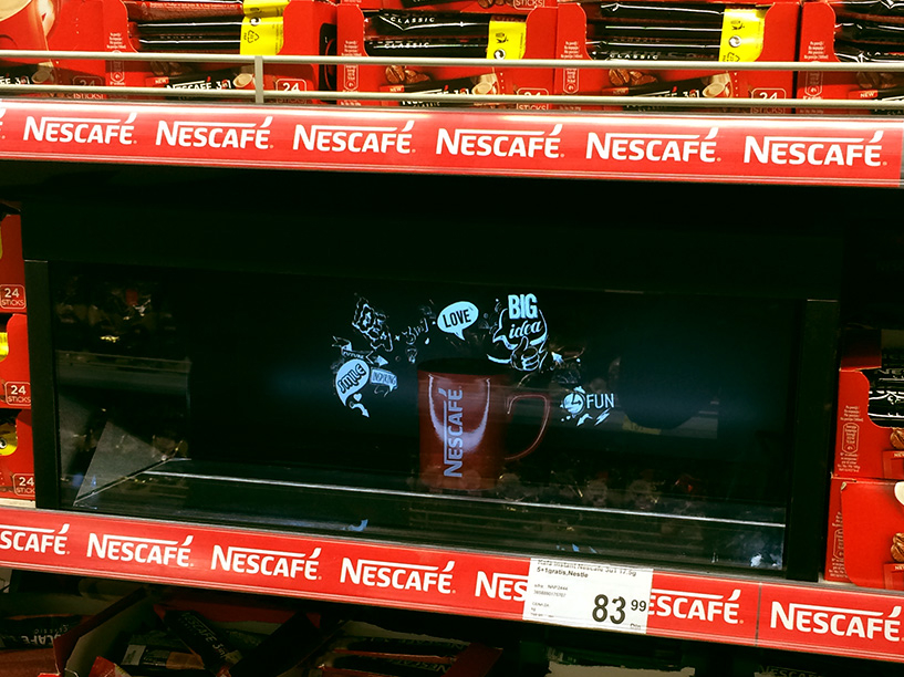 Interaktivni digitalni displej na polici u prodavnici, sa virtuelnim modelom NESCAFÉ kafe u tegli okruženim fizičkim pakovanjima proizvoda, demonstrirajući kombinaciju fizičkog i proširenog marketinga.